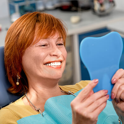 A middle-aged redhead woman looking in a mirror and smiling at the Palms Dentistry office.